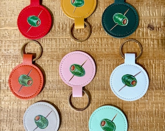 Puede incluir: Llaveros con diseño circular que muestran una aceituna verde ensartada en un palillo. Los llaveros están disponibles en varios colores: rojo, amarillo, verde, naranja, rosa, blanco, gris y verde menta. Cada llavero tiene un anillo dorado.