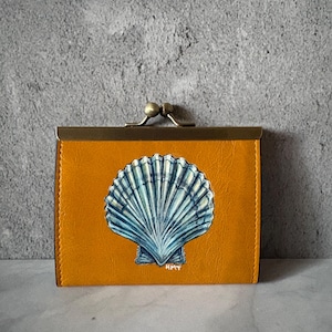 May include: Mustard yellow wallet with a hand-painted blue and white seashell design. The wallet has a brass clasp and a rectangular shape. The seashell art is centered on the front of the wallet. The artist's initials, HMT, are visible.
