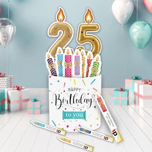 Pode incluir: Decoração de aniversário com um recipiente branco com a frase "Happy Birthday to you". Inclui o número dourado 25, velas coloridas e confetes. Várias lembrancinhas em forma de vela estão espalhadas na frente.