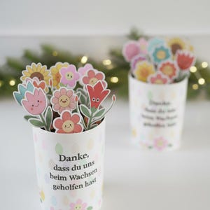 May include: Two white paper cups filled with colorful flower cutouts. The flowers have smiling faces and are in shades of pink, yellow, and red. The cups have text in German and are decorated with pastel-colored floral patterns.