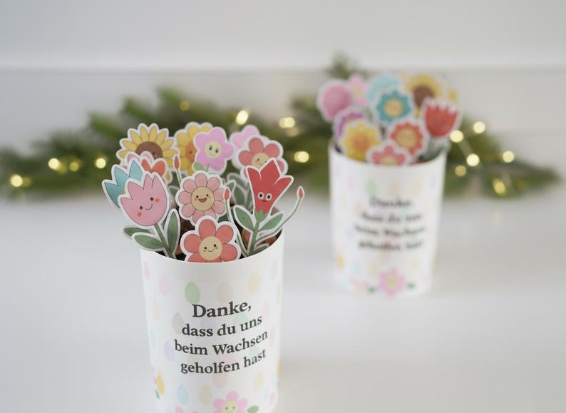 May include: Two white paper cups filled with colorful flower cutouts. The flowers have smiling faces and are in shades of pink, yellow, and red. The cups have text in German and are decorated with pastel-colored floral patterns.
