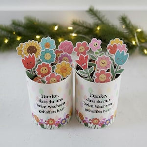 May include: Two white paper cups decorated with colorful flower stickers. Each cup is filled with various flower-shaped cutouts. The cups have text in German and are decorated with a floral border.