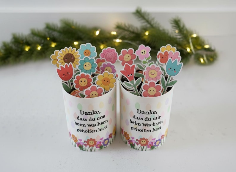 May include: Two white paper cups decorated with colorful flower stickers. Each cup is filled with various flower-shaped cutouts. The cups have text in German and are decorated with a floral border.