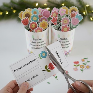 May include: Two white cylindrical containers filled with colorful paper flower cutouts. The containers have text in German. A pair of scissors cuts a card with flower graphics and German text. Decorative floral elements are scattered around.