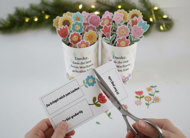 May include: Two white cylindrical containers filled with colorful paper flower cutouts. The containers have text in German. A pair of scissors cuts a card with flower graphics and German text. Decorative floral elements are scattered around.