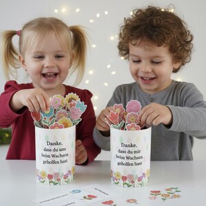May include: Two children smiling and holding flower-shaped cards. Each child is holding a white cylinder decorated with flowers and text in German. The text reads "Danke, dass du uns beim Wachsen geholfen hast."