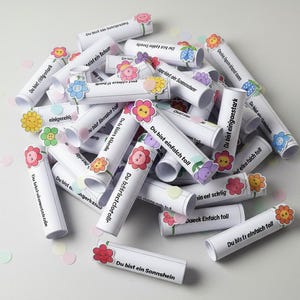May include: A collection of white paper scrolls with German text, decorated with colorful flower stickers. The scrolls are scattered on a white surface, with confetti in pastel colors. The text on the scrolls is visible.