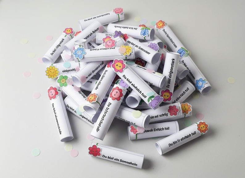 May include: A collection of white paper scrolls with German text, decorated with colorful flower stickers. The scrolls are scattered on a white surface, with confetti in pastel colors. The text on the scrolls is visible.