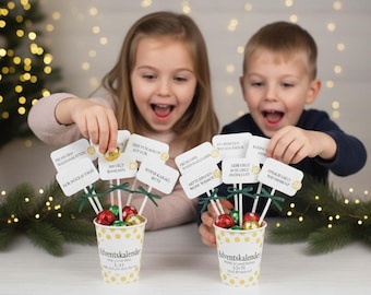 May include: Two children excitedly hold up Advent calendar treats. Each cup contains foil-wrapped chocolates and small signs with German text. The cups are decorated with polka dots and the text "Adventskalender". Festive holiday decor is in the background.