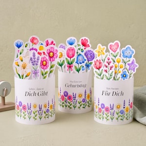 Puede incluir: Tres recipientes cilíndricos blancos decorados con ilustraciones coloridas de flores y plantas sonrientes. Cada recipiente presenta texto en alemán: 'Schön, dass es Dich Gibt', 'Alles Gute zum GEBURTSTAG' y 'Von Herzen Für Dich'.