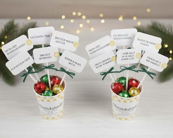 May include: Two Advent calendar cups filled with foil-wrapped chocolates in red, green, and gold. Each cup has white signs on sticks with German text and green bows. The cups have a yellow polka dot pattern.