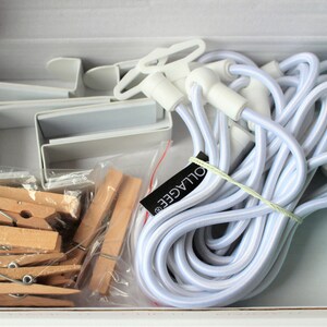 May include: A white box containing a set of white plastic picture hanging clips, white elastic cord, and wooden clothespins. The box also includes a label that says "OULAGEE".