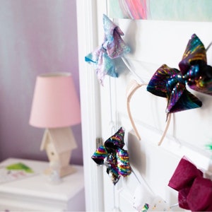 May include: A white door with a variety of colorful hair bows and headbands hanging on hooks. The bows are made of sequins, fabric, and ribbon. The headbands are made of metal and plastic.