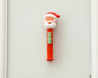 May include: A red and white Santa Claus Pez dispenser in a white frame. The dispenser has a green label with the text "Pringles" on it. The text "Kringle's Pringles" is written below the frame. The text "J. Shaw-Smith, 2013" is written in the bottom right corner of the frame.