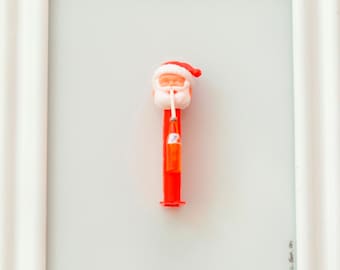 May include: A red and white Santa Claus figurine holding a bottle of soda in a white picture frame. The text "Santa + Soda" is written in the bottom left corner of the frame and "J. Henderson 2013" is written in the bottom right corner.