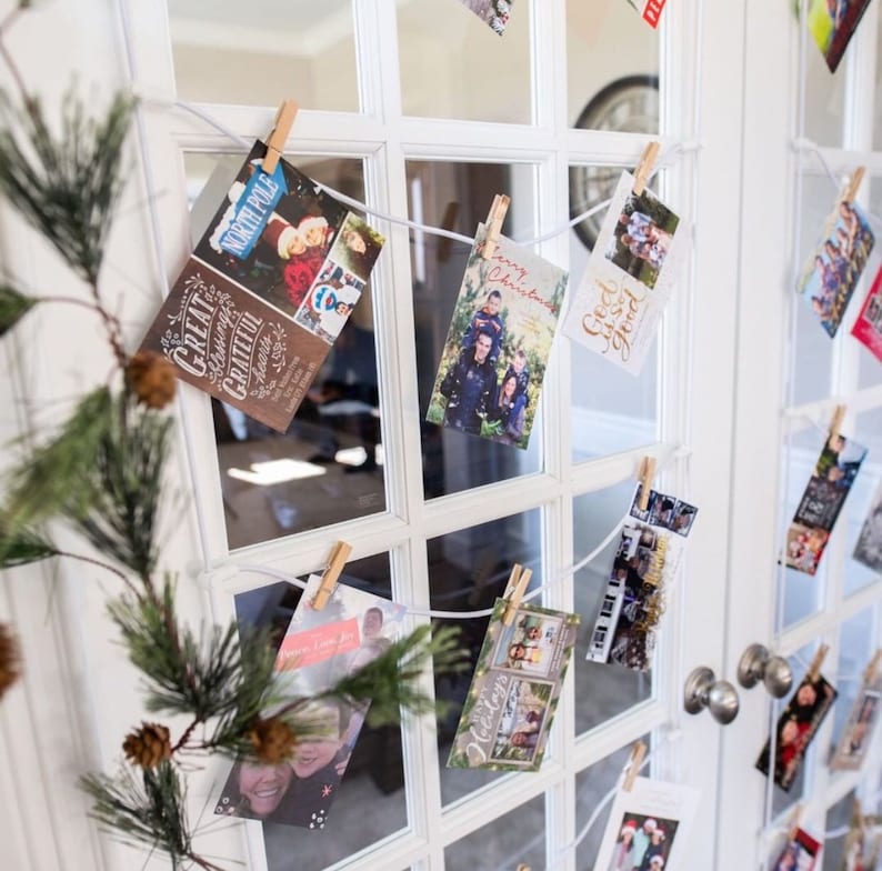 May include: A white door with a glass pane is decorated with holiday cards. The cards are hung on a white string with clothespins. The cards feature various holiday greetings and images of families.