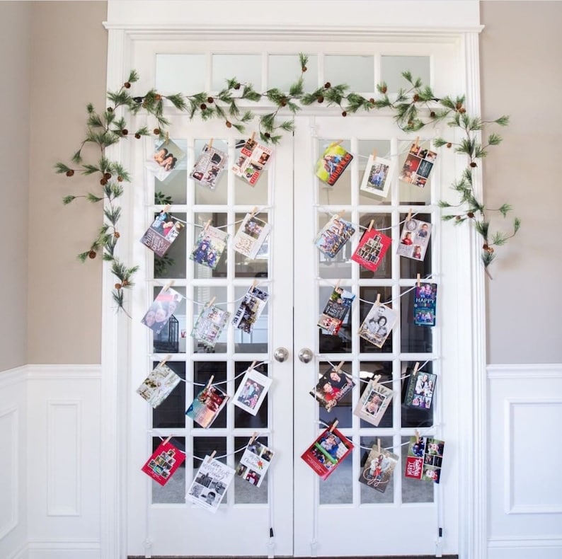 May include: A white double door with a garland of pine branches and pine cones. The garland is decorated with Christmas cards held in place with clothespins.