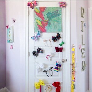 May include: A white door with a hummingbird painting and a variety of colorful hair bows and headbands hanging on it. The door is decorated with a white string and clips to hold the accessories. The name "RILEY" is spelled out in large wooden letters on the wall to the right of the door.