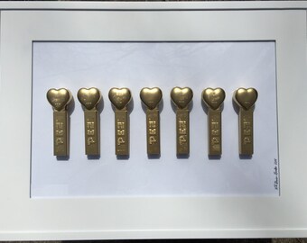 May include: A framed display of eight gold-colored heart-shaped Pez dispensers. The dispensers are arranged in a row and are all facing the same direction. The frame is white and has a white mat.