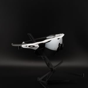 May include: White and black sports sunglasses with a modern design. The sunglasses have a white frame with black detailing and dark lenses. The image is set against a dark background, highlighting the sunglasses' design.