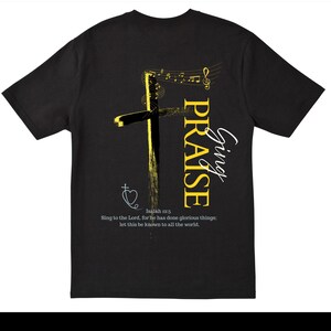 Puede incluir: Camiseta negra con un diseño de cruz amarilla, notas musicales y las palabras "Sing Praise" en blanco y amarillo. La camiseta incluye un versículo bíblico de Isaías 12:5.