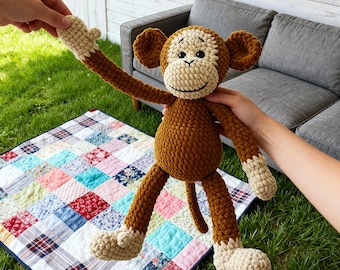 May include: A handmade crochet monkey doll in shades of brown and beige. The monkey has long arms and legs, with a smiling face. The doll is held up against a backdrop of green grass and a colorful patchwork quilt.