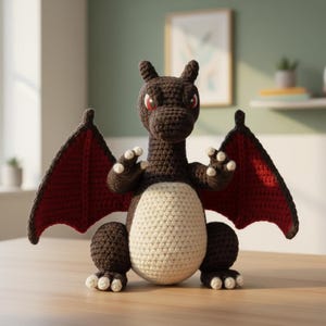 Puede incluir: Una figura de dragón de ganchillo con cuerpo marrón, alas rojas y vientre color crema. El dragón tiene garras blancas y ojos rojos. Las alas son de un rojo intenso con borde marrón oscuro. El dragón está sobre una superficie de madera.