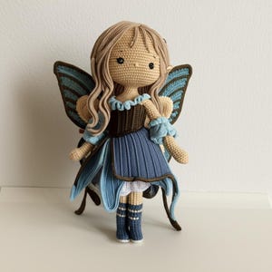 May include: A handmade crochet doll in the shape of a fairy. The doll has long, wavy, light brown hair and large blue and brown wings. She wears a blue dress with a brown bodice and blue accents. The doll is approximately 10 inches tall.