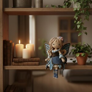 May include: A handmade crochet fairy doll with long, light brown hair and blue butterfly wings. The doll wears a blue dress with a dark brown bodice and is adorned with a flower crown. The doll is displayed on a wooden shelf with lit candles and books.