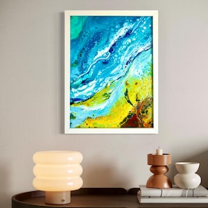 Può includere: Stampa artistica astratta in una cornice bianca. L'opera presenta un vivace mix di blu, turchese, giallo e bianco, creando un design fluido a forma di onda. Il dipinto è esposto sopra un tavolo con una lampada e oggetti decorativi.