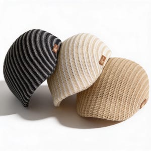 May include: Three knitted hats in a studio setting. The hats are in black and white, cream and white, and tan. Each hat has a small brown label. The hats are arranged on a white background.