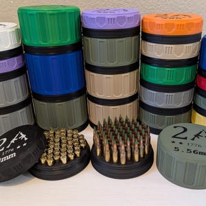 May include: A collection of colorful, cylindrical containers with ribbed sides and flat lids. Some lids display text and symbols, including "2A" and caliber designations like "9mm" and "5.56mm". The containers are stacked and arranged with ammunition.