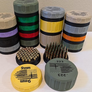 May include: A collection of colorful ammunition containers and rounds. The containers are stacked in various colors, including green, gray, yellow, and black. The lids are labeled with text and a flag design. The image also shows a collection of bullets.