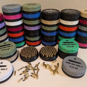 May include: A collection of colorful ammunition storage containers with the text "COME AND TAKE IT" and caliber designations like .380, 45acp, and 22LR. The containers are stacked in various colors, and some are filled with bullets.