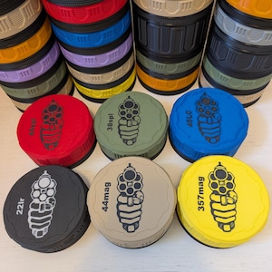 May include: A collection of colorful, stacked containers with black and tan accents. The lids are red, green, blue, black, tan, and yellow, each labeled with a caliber designation and a gun graphic.