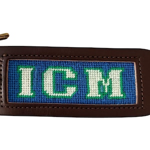 May include: A brown leather keychain with a blue and green embroidered panel. The panel displays the initials "ICM" in white thread. Gold-colored hardware and a key are attached.