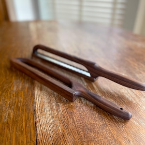 May include: Two wooden vegetable peelers with a dark brown finish are displayed on a wooden table. The peelers have a rectangular handle with a cutout and a stainless steel blade. The blades are visible in the center of the peelers.