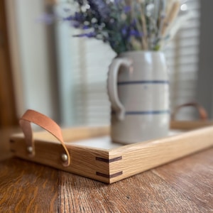 May include: A rectangular wooden serving tray featuring light-colored wood grain, dark wood corner splines, and two tan leather handles secured with metal rivets. A ceramic pitcher with blue horizontal stripes sits inside the tray.