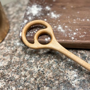 May include: A light-colored wooden kitchen utensil featuring a long handle and a head designed with two circular cutouts of different sizes. The smooth, natural wood grain is visible throughout the entire length of the stirring tool.