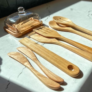 May include: A collection of wooden kitchen utensils, including spoons, spatulas, and butter knives, alongside a wooden butter dish with a glass lid. The items are arranged on a white countertop, bathed in natural light.