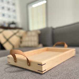 May include: A rectangular wooden serving tray with light-colored wood and leather handles. The tray has a simple design with visible joinery at the corners. The leather handles are attached with brass-colored rivets. The tray is designed for serving food or drinks.