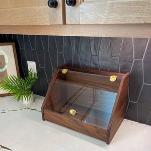 May include: A wooden display case with a clear glass front and top. The case is made of dark brown wood and has brass-colored hardware. The case is angled and sits on a white countertop.