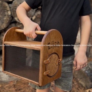 May include: A wooden bug catcher with a handle and a mesh front. The catcher has a small door and the name "Elijah" is engraved on the side. The bug catcher is held by a person.
