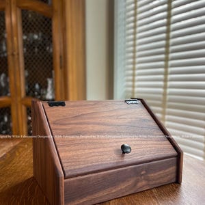 May include: A handcrafted wooden box with a slanted lid, featuring a dark brown finish. The box has a black knob and hinges, and is displayed on a wooden surface. The text "Designed by Wilde Fabrications" is visible.