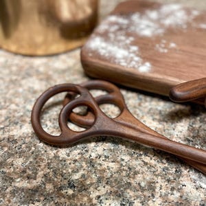 May include: A pair of dark brown wooden dough blenders with circular handles and a unique design. A wooden cutting board with flour and a copper container are in the background. Kitchen tools.