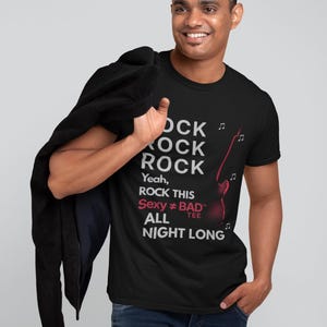 Puede incluir: Camiseta negra con el texto "ROCK ROCK ROCK" y "ROCK THIS Sexy ≠ BAD ALL NIGHT LONG" en blanco y rojo. Un gráfico de guitarra roja está en el lado derecho. La persona lleva vaqueros azules.