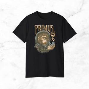 May include: Black t-shirt featuring a detailed graphic of a monkey in a space helmet smoking a pipe. The word "PRIMUS" is printed above the illustration in a vintage style font. The design is in shades of brown, tan, and gold.