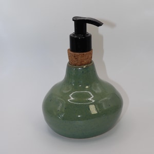 May include: Green ceramic soap dispenser with a black pump and cork stopper. The dispenser has a rounded, bulbous shape and a glossy finish. The pump is made of black plastic.