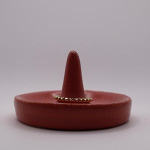 May include: A terracotta-colored ring holder with a central cone and a shallow dish base. A gold ring with a geometric design rests on the holder. The ring holder is on a white surface.