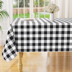 May include: A black, white, and gray checkered tablecloth covers a wooden table. The tablecloth is draped over the table, with a portion hanging off the side. A vase of white flowers sits on the table, along with a plate and glasses.